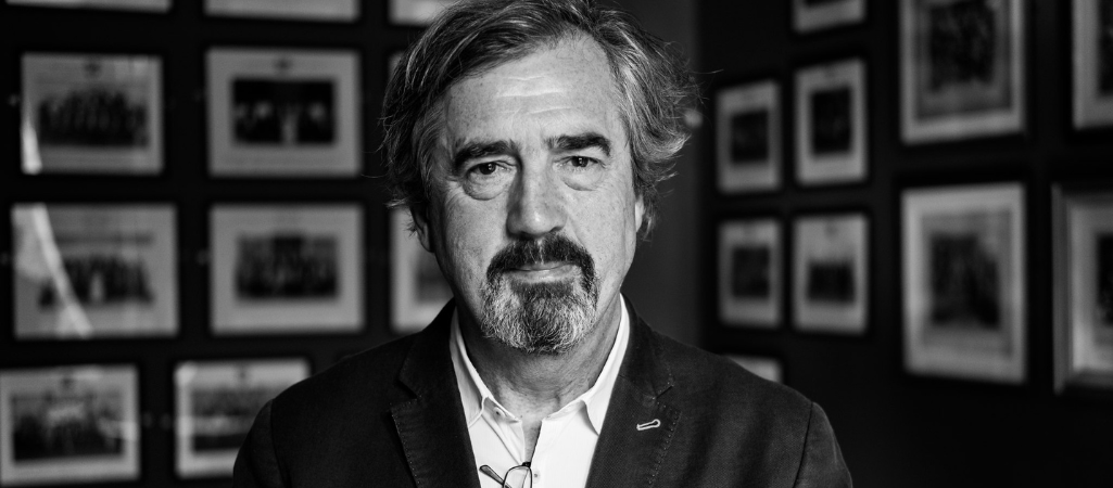 Irlandzki pisarz Sebastian Barry. Czarno-białe zdjęcie mężczyzny w średnim wieku z siwiejącymi włosami i brodą, ubranego w marynarkę i koszulę, stojącego na tle ściany z licznymi oprawionymi fotografiami (fot. Chris Boland / www.chrisboland.com CC BY-NC-ND 2.0)